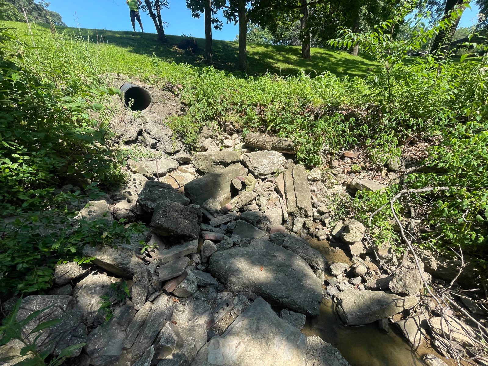 Failing pond dam outlet near Decatur, Illinois — the pipe daylights part way down the downstream face and the flow has cut a gully through broken-concrete riprap below it